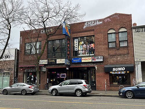 A row of ground floor shops in an aging brick building on Broadway, cars parked along the curb and graffiti tags on the upper brick.