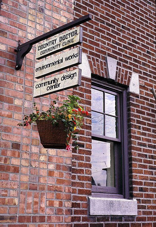 A brick building corner with an iron sign bracket, hanging signs, a paned window with a granite lintel, and a flower box mounted below the sill.