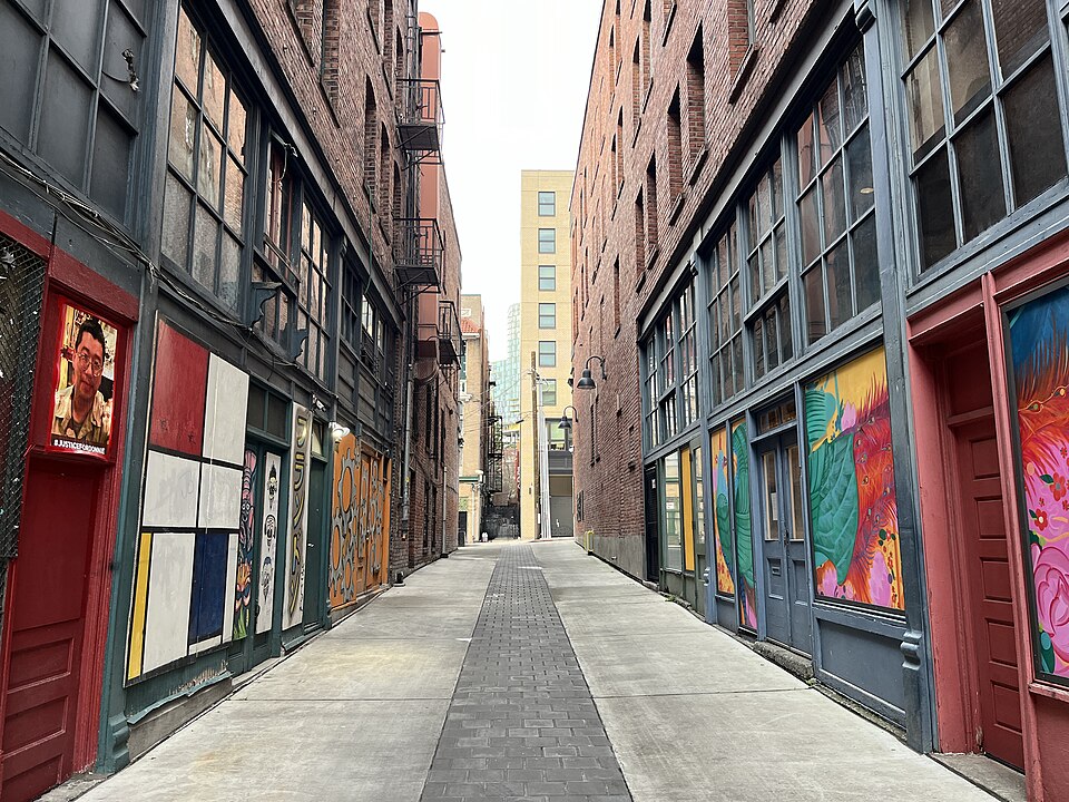 A plain stretch of Canton Alley, brick walls rising four storeys on both sides with a fire escape and vents, chain link fencing near the far end.