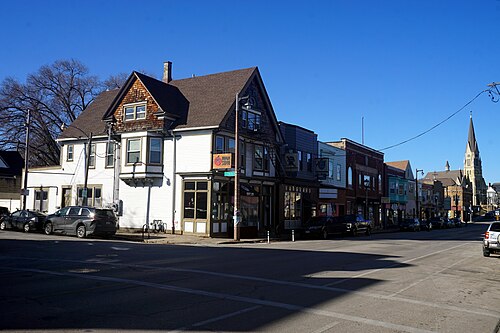 A wide corner on South Kinnickinnic Avenue, wood shingle and brick storefronts under a shared roofline with a church spire down the block.