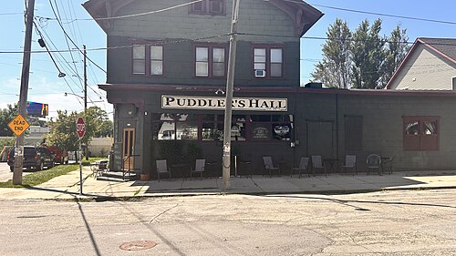 Puddler's Hall, a two storey dark green wood building with a painted sign reading Puddler's Hall Est 1873 over the door and chairs out front.