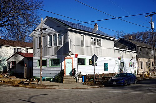 The Cactus Club, a two storey frame venue building in grey and white siding on a street corner, solar panels on the roof and picnic tables out front.