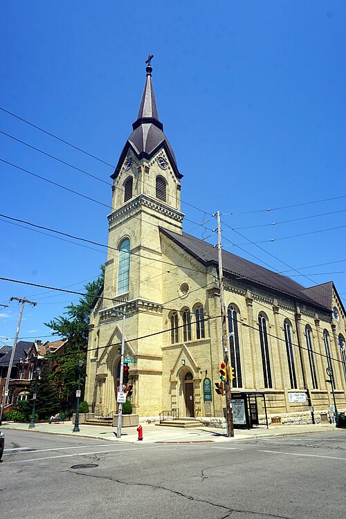 St. Hedwig's Church from the corner, yellow brick with a tall clock tower and steeple, arched stained glass windows along the nave, a Brady Street sign visible on the corner post.