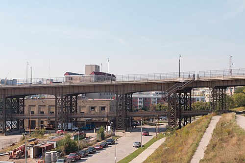 The Holton Street Viaduct from below, its steel girders on concrete piers with the timber footbridge deck slung underneath, industrial buildings and a rail cut beneath.