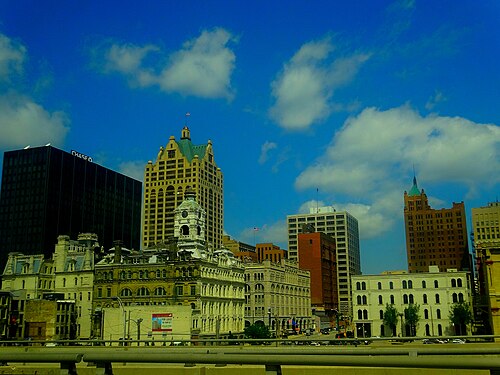 A view from a bridge of the skyline, a dark glass tower beside a tan stone building with a domed green roof, older masonry storefronts in the foreground.