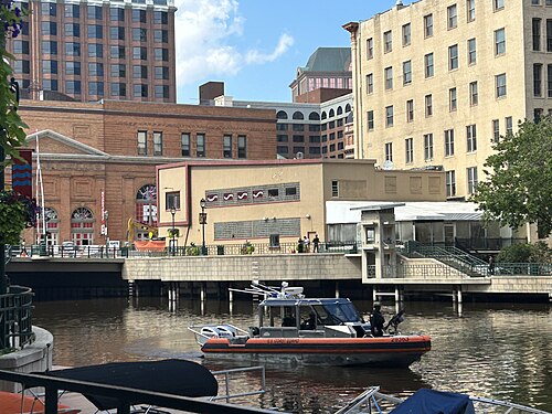 A boat on the river below a tan brick building with tall arched windows, a black glass office tower and a red brick warehouse behind it.