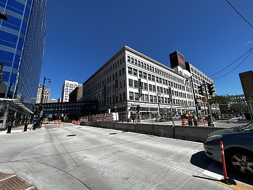 A wide downtown street corner, stone faced office buildings and a modern black tower, orange traffic barrels along the curb.