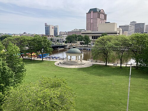An open lawn along the river with a domed white pavilion at its edge, paddle boats moored at the bank and office towers rising behind the trees.