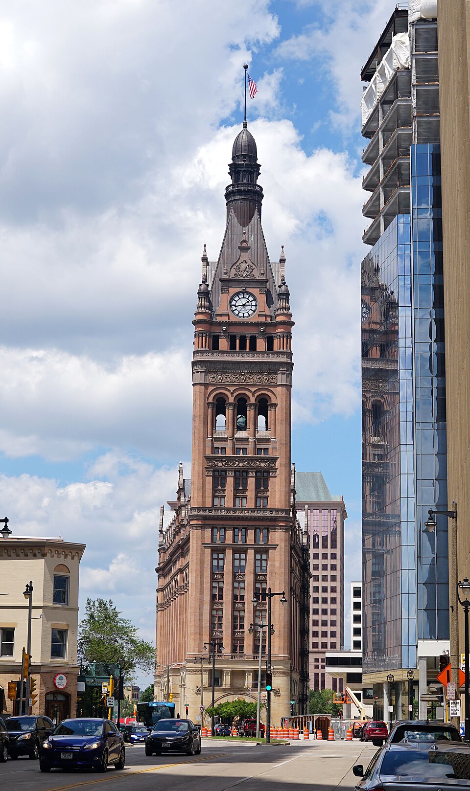 Milwaukee City Hall's brick and terra cotta tower rising above the street, the clock face and copper spire at the top, construction barriers and cars along Wells Street below.