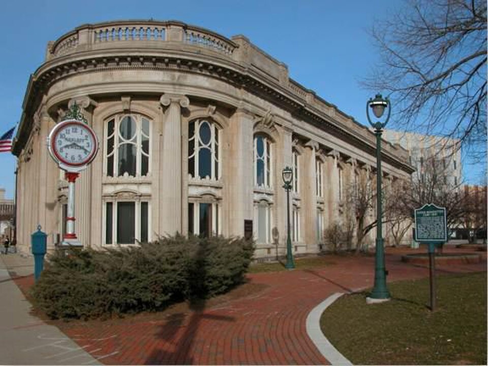 The former Second Ward Savings Bank building's curved corner, pale stone with paired arched windows and a fluted cornice, a sidewalk clock mounted on a post out front.