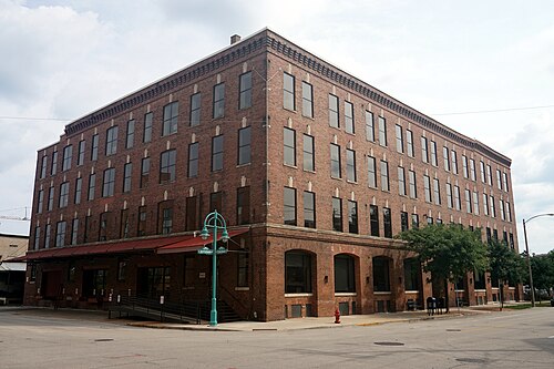 A five storey brick building with a full grid of factory windows and a tall brick smokestack rising at the corner, traffic signals and street signs at the intersection below.
