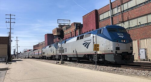 Amtrak's Empire Builder passing a long multistory red brick industrial building on the tracks, an old rooftop sign above the cornice.