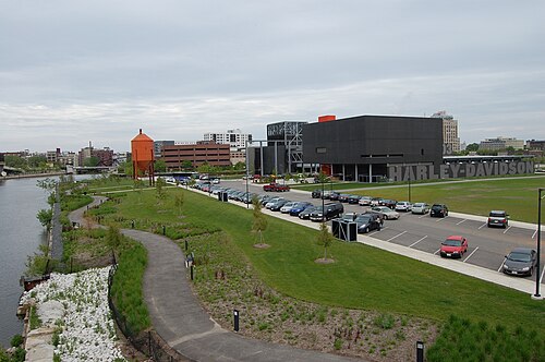 An elevated view of the Harley-Davidson Museum grounds on the Menomonee River, an orange steel water tower on the bank and downtown's towers across the water.