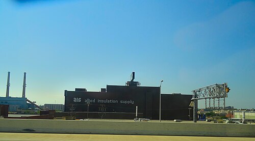 Allied Insulation Supply's dark metal warehouse along the freeway edge on 12th Street, its name painted across the wall and smokestacks rising behind it.