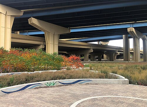 Concrete freeway viaduct piers on tapered legs over a landscaped plaza, native plantings along a paved path underneath.