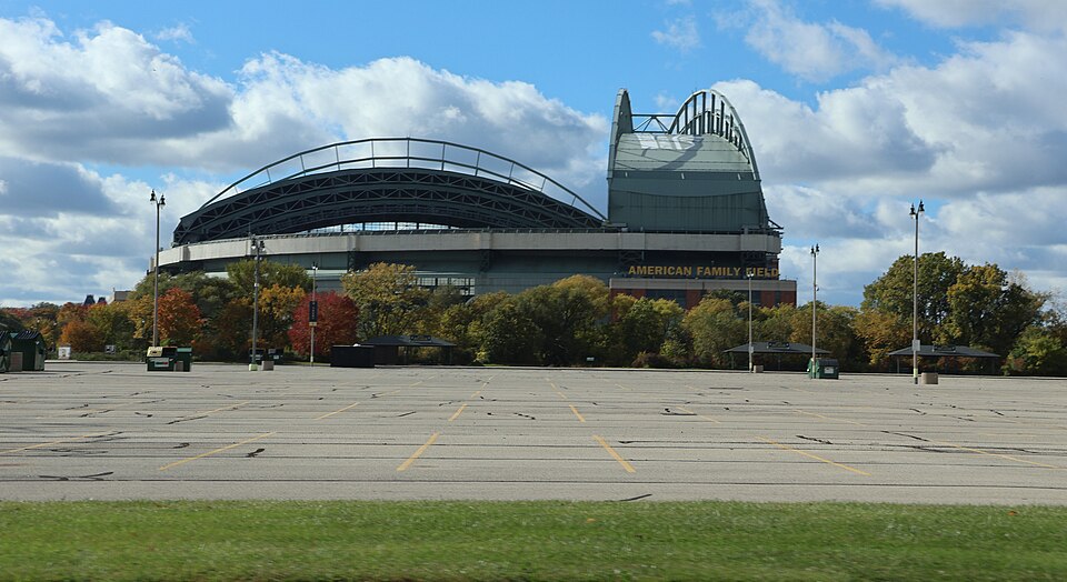 American Family Field's retractable roof and fan shaped steel arches behind an empty parking lot, the stadium's name across the wall.