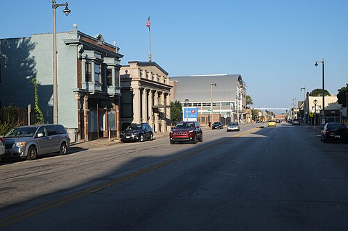 National Avenue's commercial block, a pale blue Italianate storefront and a stone bank building with columns, a curved metal roofed arena beyond and cars parked along the curb.