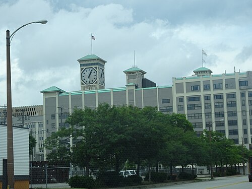 The Allen-Bradley clock tower's octagonal faces rising over a long grey factory roofline with green painted cornices and flagpoles, trees along the street in front.