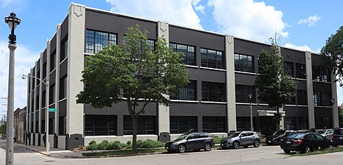 The Junior House building, a three storey dark grey stuccoed industrial block with tall steel frame windows set in cream pilaster bays, street trees along the sidewalk.
