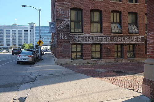 A red brick warehouse corner with a painted Schaefer Brushes ghost sign on the wall, arched window heads and a brick paved apron at the sidewalk.