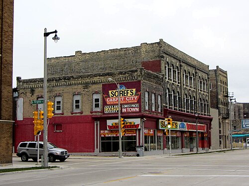 A cream brick corner building with a red painted ground floor storefront and a carpet store's wall signs, arched upper windows and a traffic light at the corner.