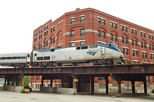 An Amtrak train passing under a red brick warehouse on a riveted steel elevated trestle, arched window bays along the building's corner.