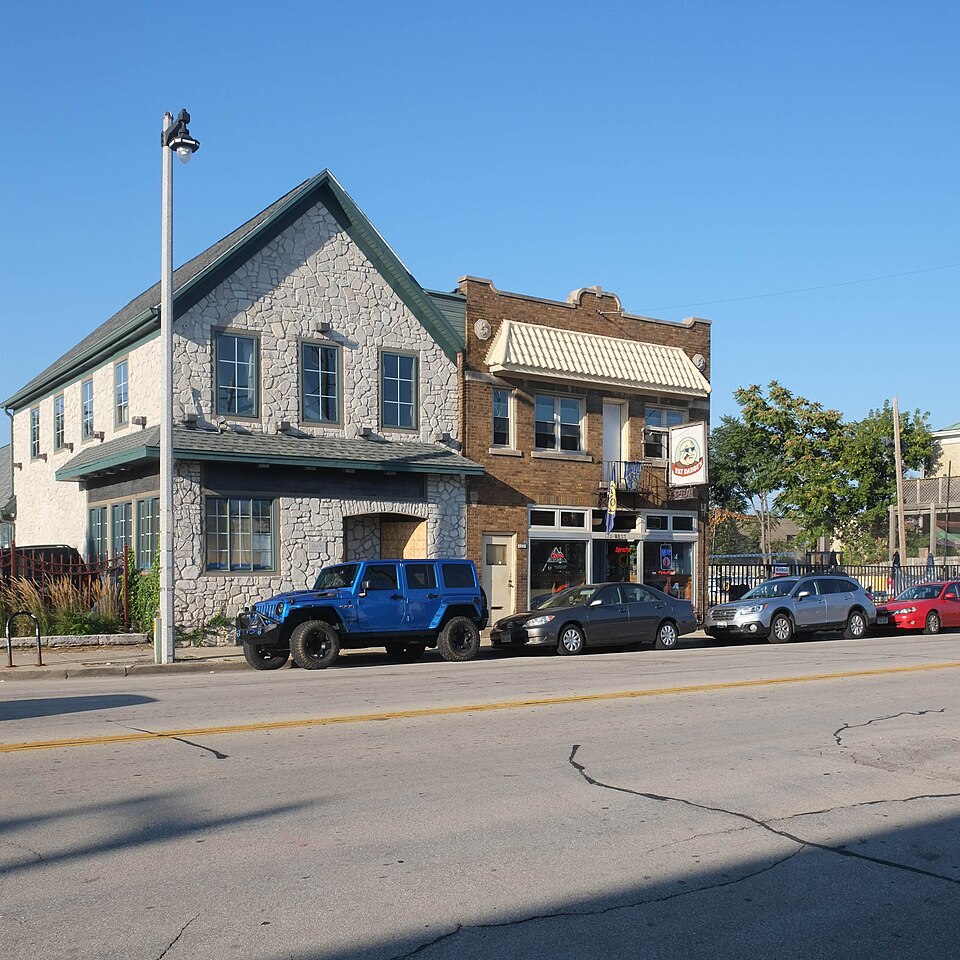 Two storey buildings on a wide street, one in grey stacked stone with a peaked green roof, the other in brick with a striped awning and the Fat Daddy's sign, a Jeep and three cars parked along the curb.